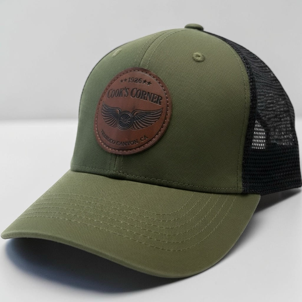 Cook's Corner olive green trucker hat with black mesh back and circular faux-leather logo patch on front panel